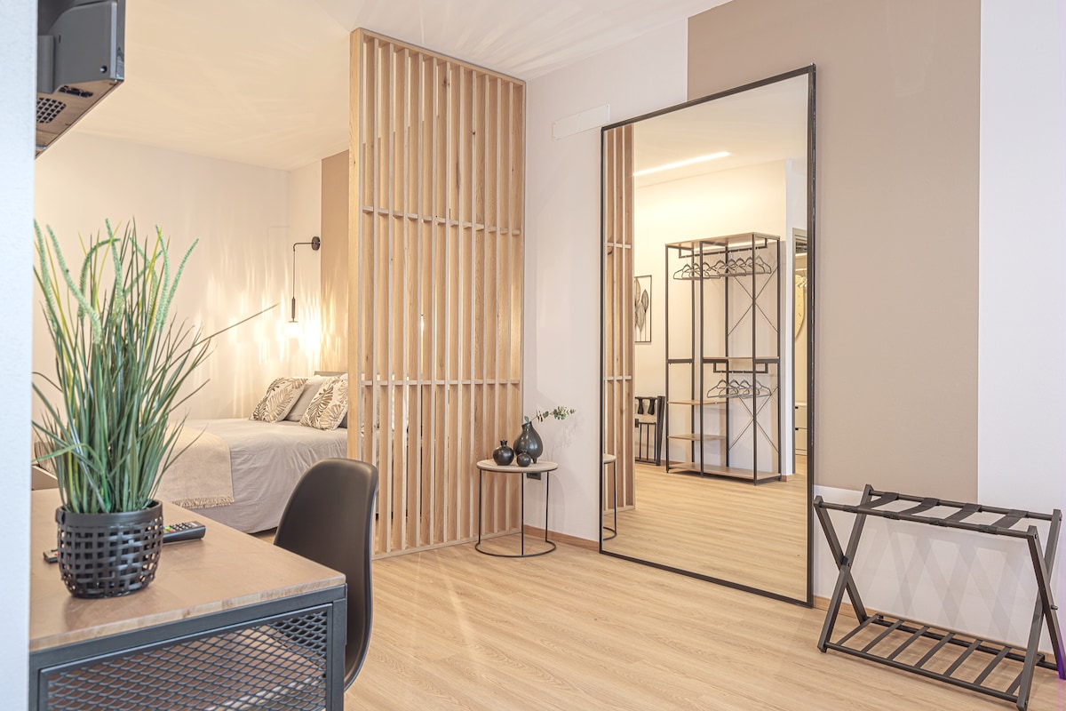A modern living space is highlighted by a spacious bed, accompanied by a geometric wooden partition. A large mirror reflects the light and enhances the sense of openness. A work desk and a foldable drying rack are positioned thoughtfully, creating a functional layout.