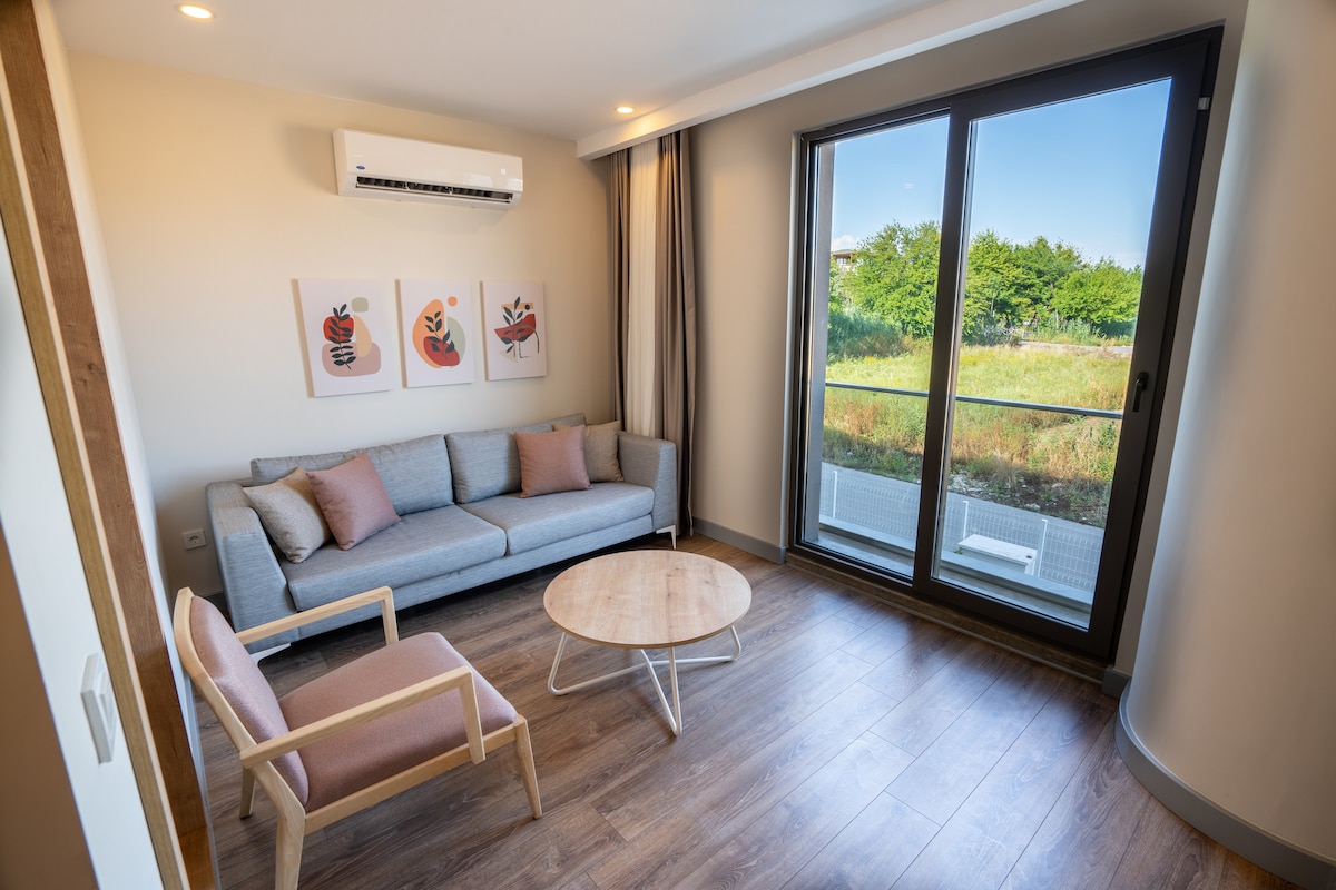 A well-lit living area is presented with large glass doors offering a view of greenery outside. A gray sofa and a pink armchair are arranged around a round wooden coffee table. Three framed art pieces add a touch of color to the light-colored walls.