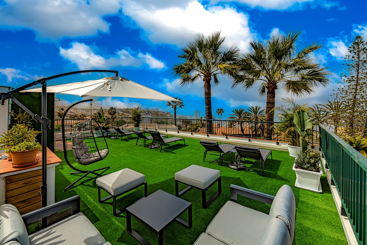 A rooftop terrace is seen, featuring several outdoor seating areas with lounge chairs and small tables arranged on green lawn-like flooring. Tall palm trees provide shade, and distant city views are visible against a bright blue sky.