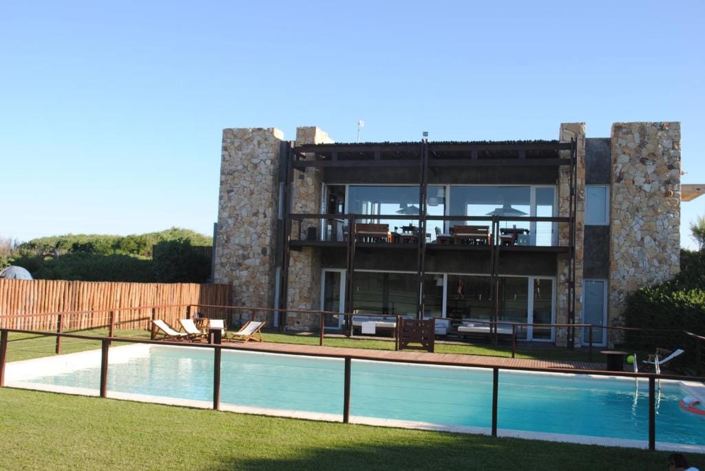 The modern structure is set against a clear blue sky, featuring large glass windows that reflect the surrounding greenery. A spacious outdoor area includes a swimming pool and lounge chairs, providing a relaxing space for enjoyment. Wooden fencing surrounds the property, enhancing privacy.