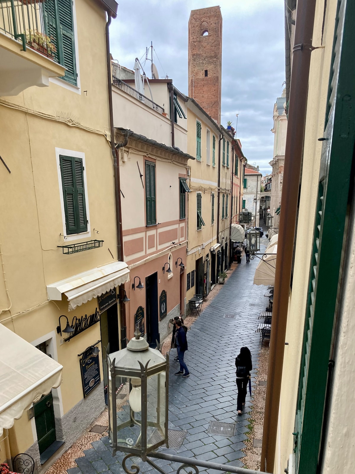 A narrow pedestrian street is depicted, showcasing pastel-colored buildings with green shutters. A tall brick tower rises prominently at the end of the street. People can be seen walking, and outdoor seating is visible under awnings, adding to the lively atmosphere.