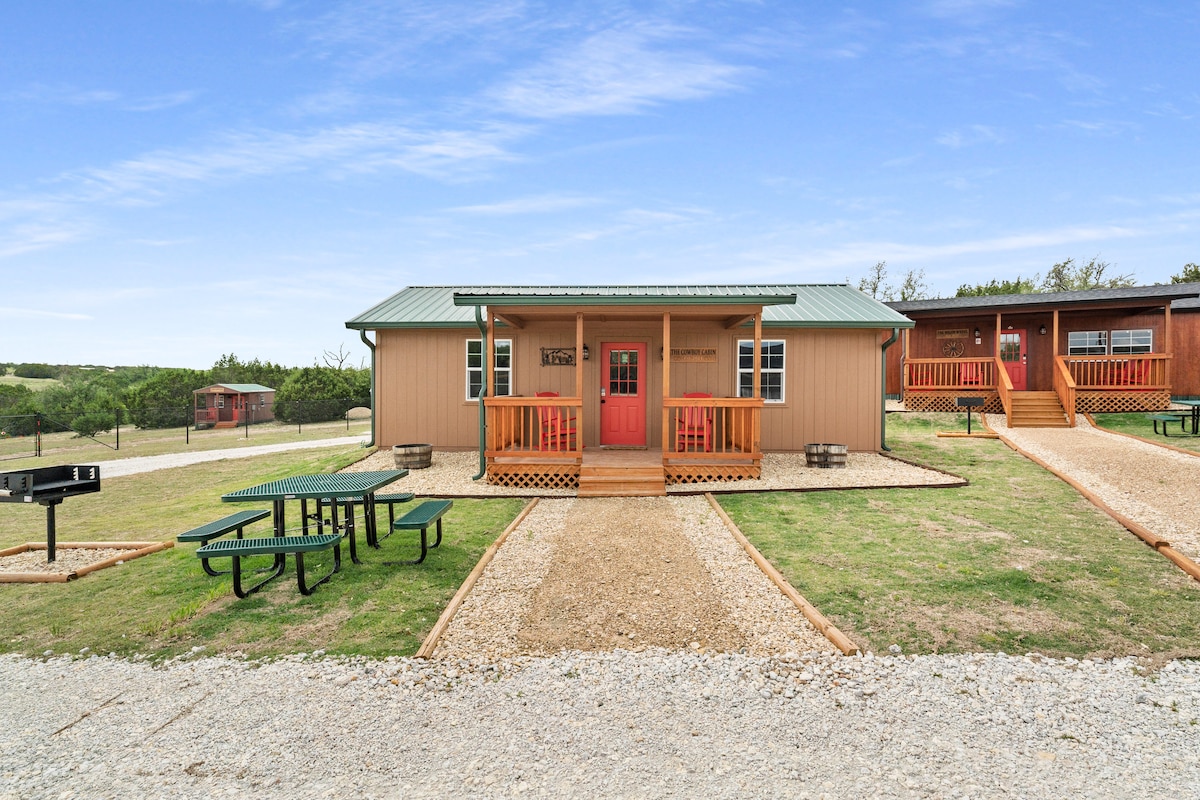 A rustic cabin is visible in a rural setting, featuring a light brown exterior and a welcoming red door. A gravel pathway leads to the entrance, surrounded by green grass and picnic tables. Barbecue grills are positioned nearby, enhancing outdoor gathering spaces.