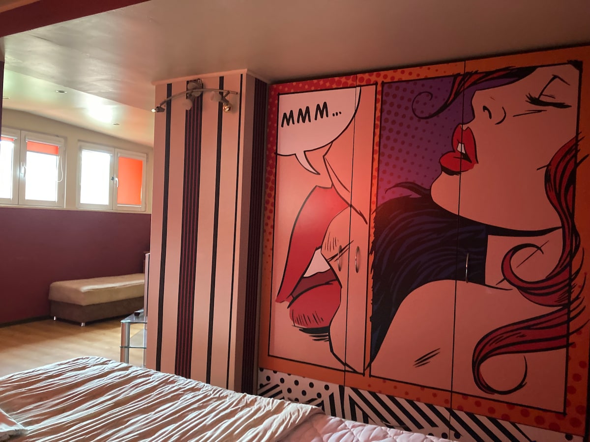 A vibrant wall mural depicting a woman's face is the focal point of the room, complemented by geometric-patterned walls. A cozy bed is placed in front, and a seating area is visible in the background, beside large windows that allow natural light to enter.