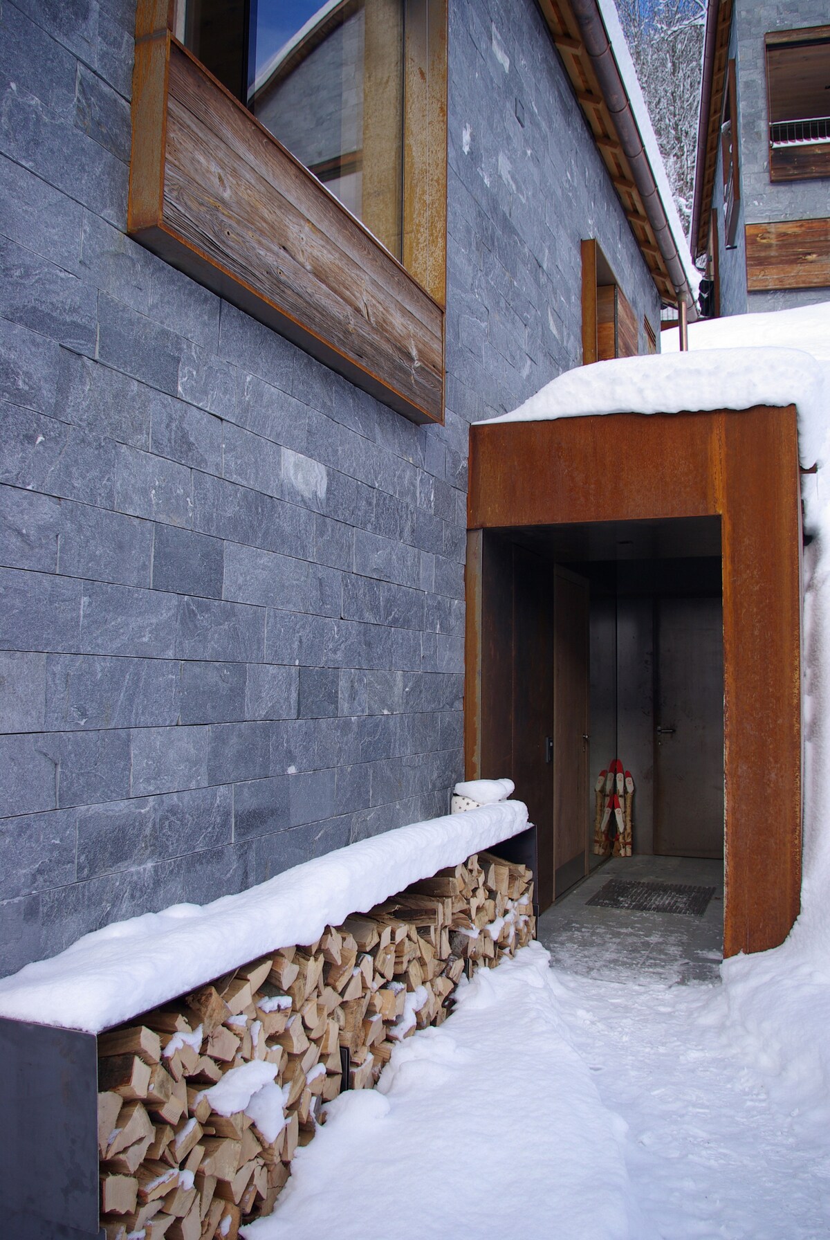 The entrance of the house is framed by a sleek wooden awning and natural stone walls, partially covered in snow. A stacked wood pile offers warmth, while a metal door leads inside. Large windows allow light to filter through, enhancing the modern architectural design.
