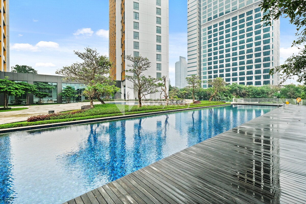 An outdoor swimming pool reflects the sky, bordered by a wooden deck. Lush greenery frames the area, enhancing the serene environment, while modern high-rise buildings stand in the background, contributing to the contemporary setting.
