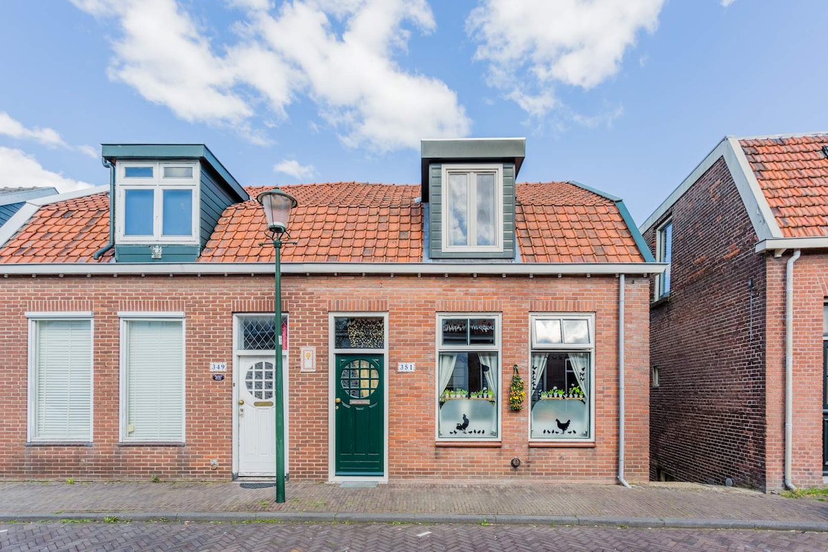 A charming brick facade features large windows adorned with floral decorations. The entry door is painted in a deep green, contrasting with the warm tones of the red roof tiles. The building has a symmetrical design, contributing to its inviting appearance.