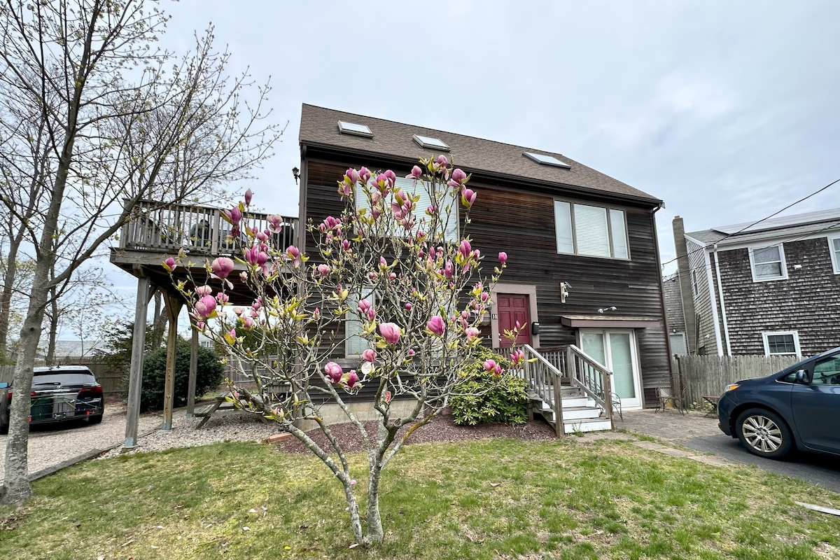 The exterior of a two-story wooden house is shown, featuring a spacious entryway and a deck. A flowering magnolia tree is visible in the foreground, with vibrant pink blossoms contrasting against the house's dark wood. A landscaped yard surrounds the property.