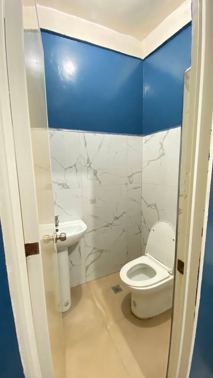 A compact bathroom is presented with blue walls and smooth, light-colored tiles. The space features a toilet and a small sink, each positioned against marble-patterned panels that enhance the clean aesthetic. Natural light is available through a discreet opening.