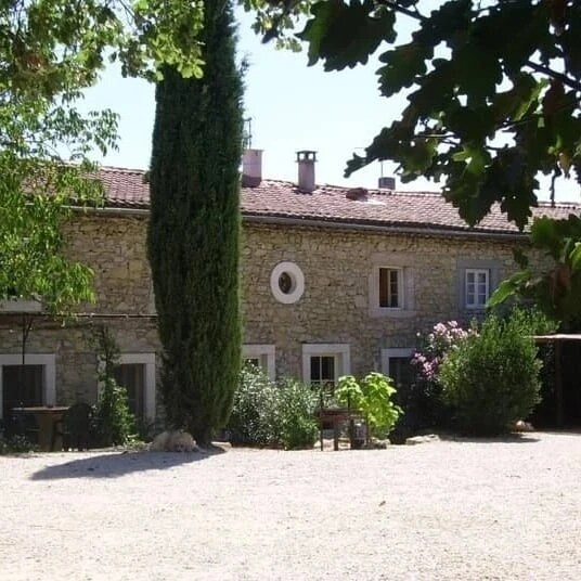 The exterior of the charming stone house features a circular window and is surrounded by lush greenery. Tall cypress trees are visible alongside flowering shrubs, enhancing the peaceful ambiance of the setting. A gravel area is located in the foreground, creating a natural pathway.