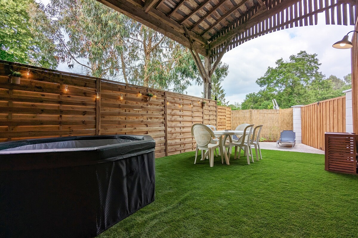 An outdoor patio area features a covered space with artificial grass. A black jacuzzi is positioned in one corner. A dining table with six white chairs is set under the structure, providing a comfortable spot for relaxation. Lush greenery is visible in the background.