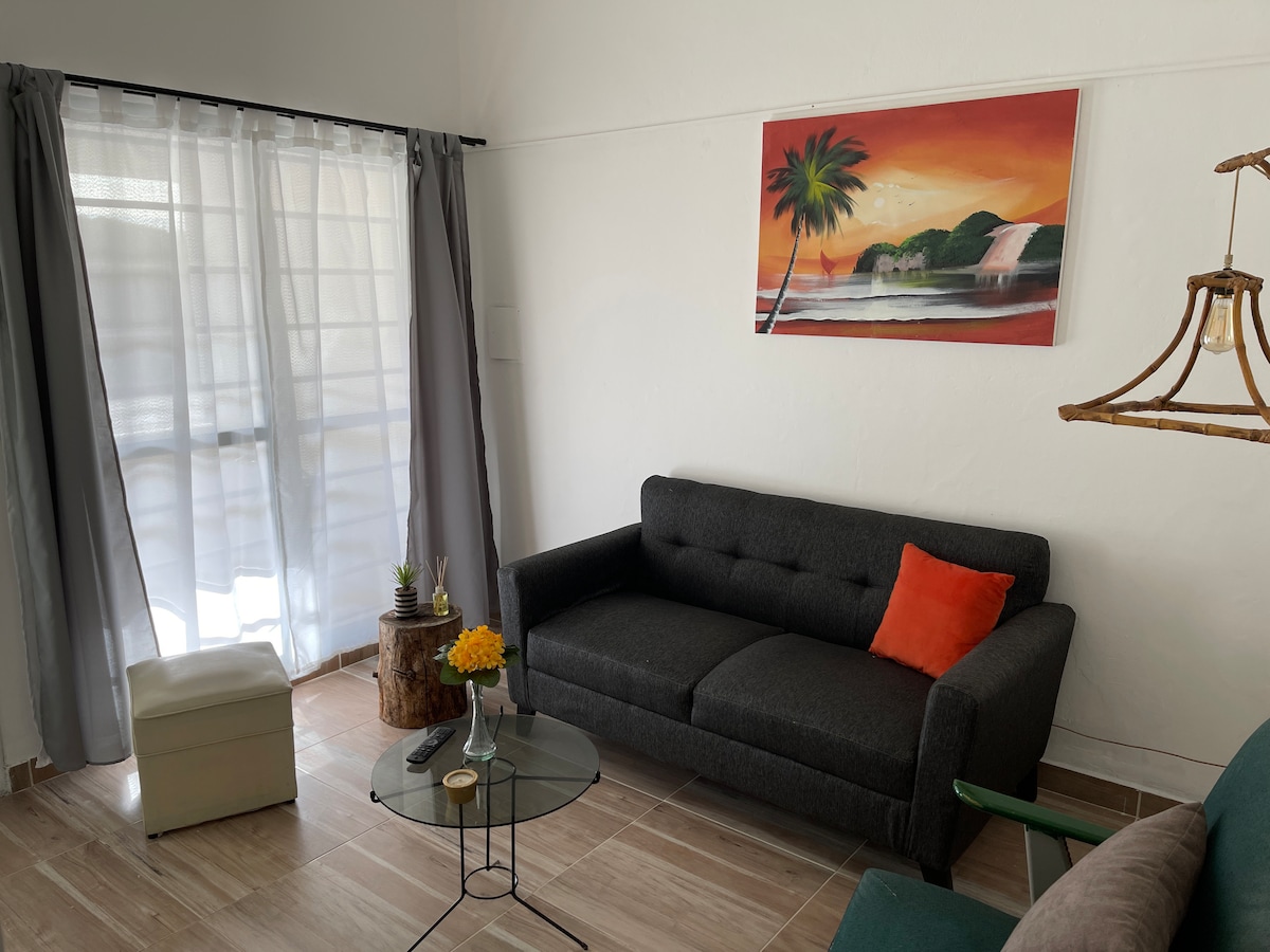 A cozy living area is highlighted by a dark gray sofa adorned with a bright orange cushion. A small glass table sits in front, accompanied by a potted plant and a wooden stump table. Natural light filters through sheer curtains, enhancing the relaxed ambiance.