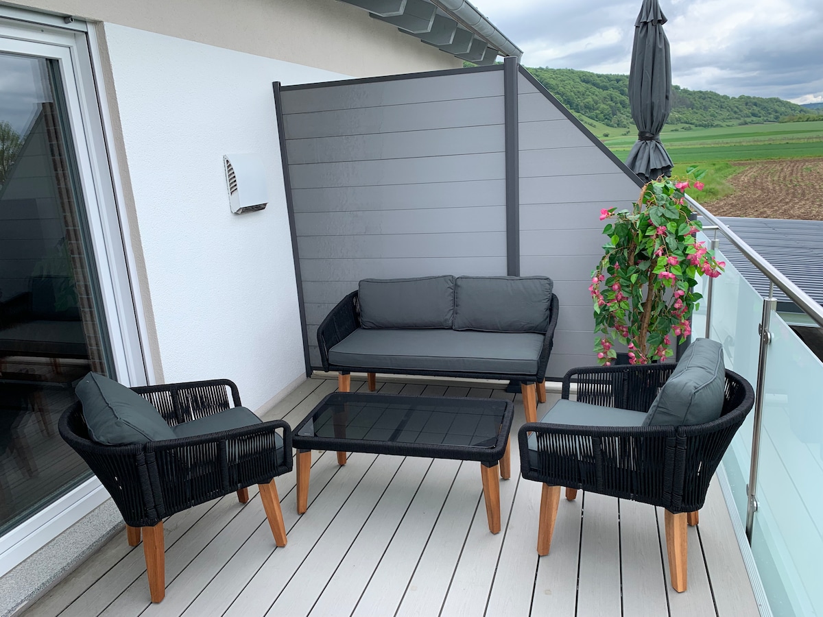 A private balcony features a comfortable seating area with a dark wicker couch flanked by two matching chairs. A glass tabletop sits at the center. Green plants and an umbrella provide a touch of nature and shade, enhancing the relaxing outdoor space.