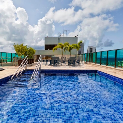 An outdoor rooftop pool is set against a backdrop of cloudy skies and city views, surrounded by a terrace with lounge chairs. The pool features a tiled design in shades of blue, with steps leading into the water.