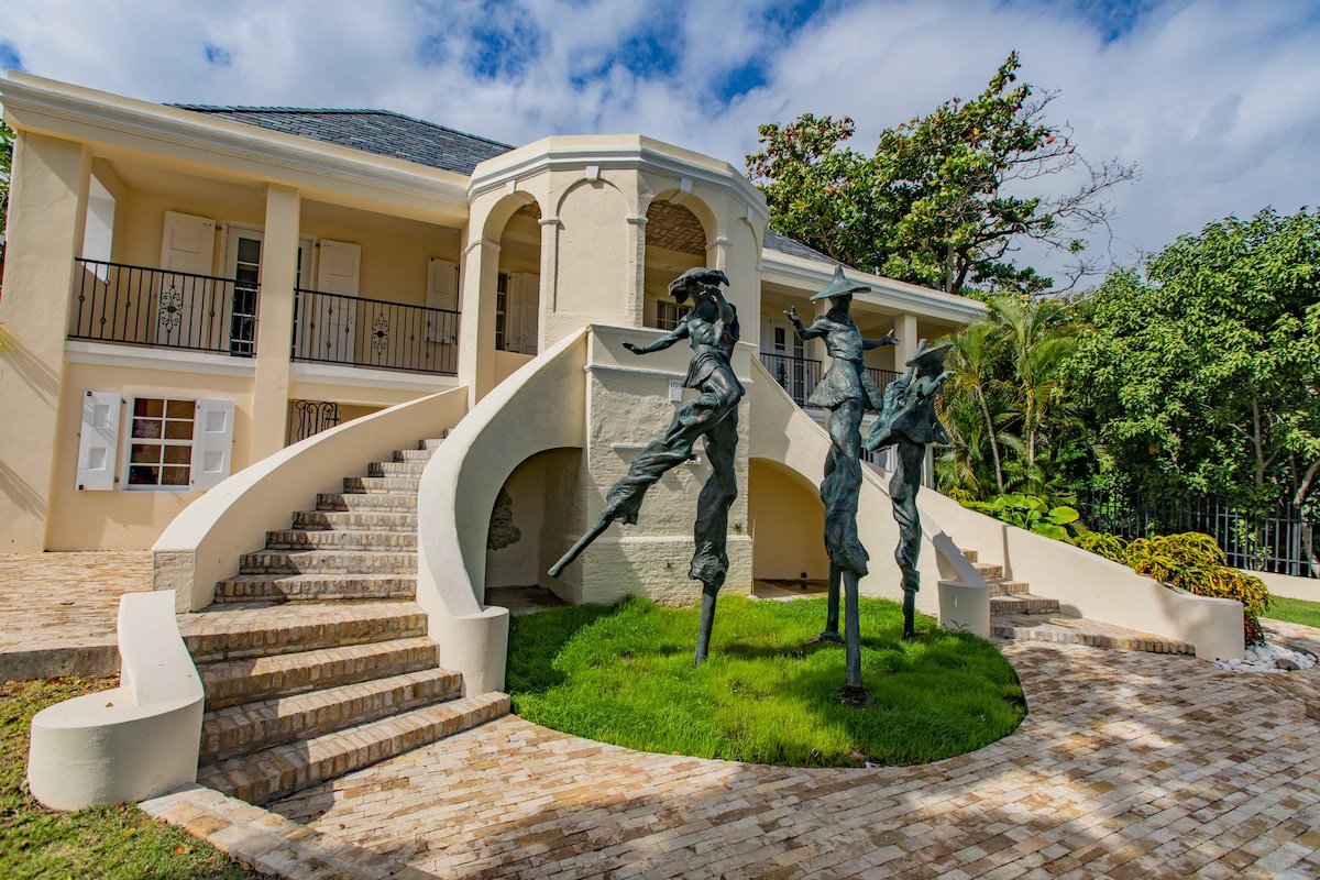 A historic building with a curved grand staircase leads to an upper gallery. In the foreground, a striking bronze sculpture features three Moko Jumbies, set amidst a well-maintained green lawn. Surrounding vegetation adds a natural touch to the inviting entrance.