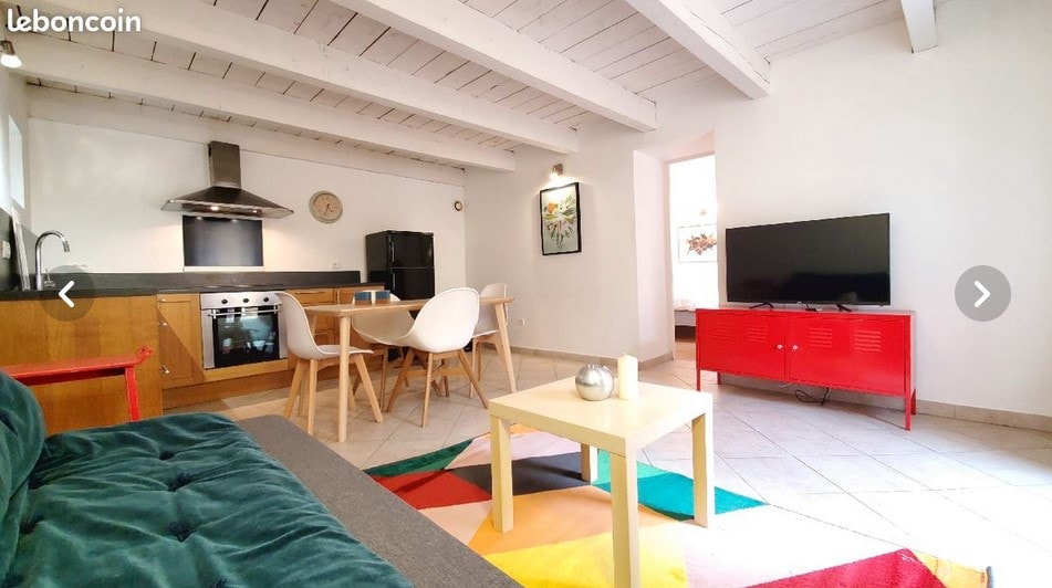 A bright living area is shown, featuring a cozy sofa in green, a light-colored dining table surrounded by white chairs, and a modern kitchen with stainless steel appliances. A television is positioned on a red cabinet. Natural light fills the space through windows with light walls.