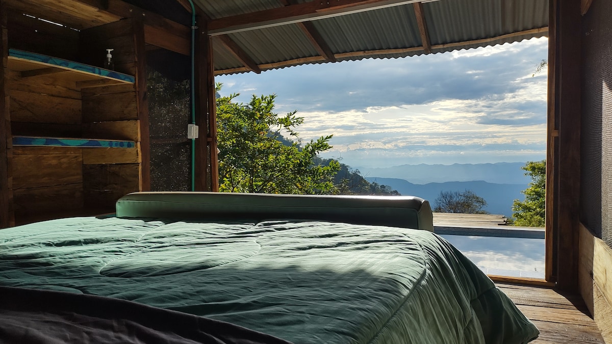 A spacious bed with a green coverlet is positioned beside large wooden windows, providing an unobstructed view of the distant mountains and sky. Natural light floods the interior, enhancing the earthy wooden textures of the cabin walls.