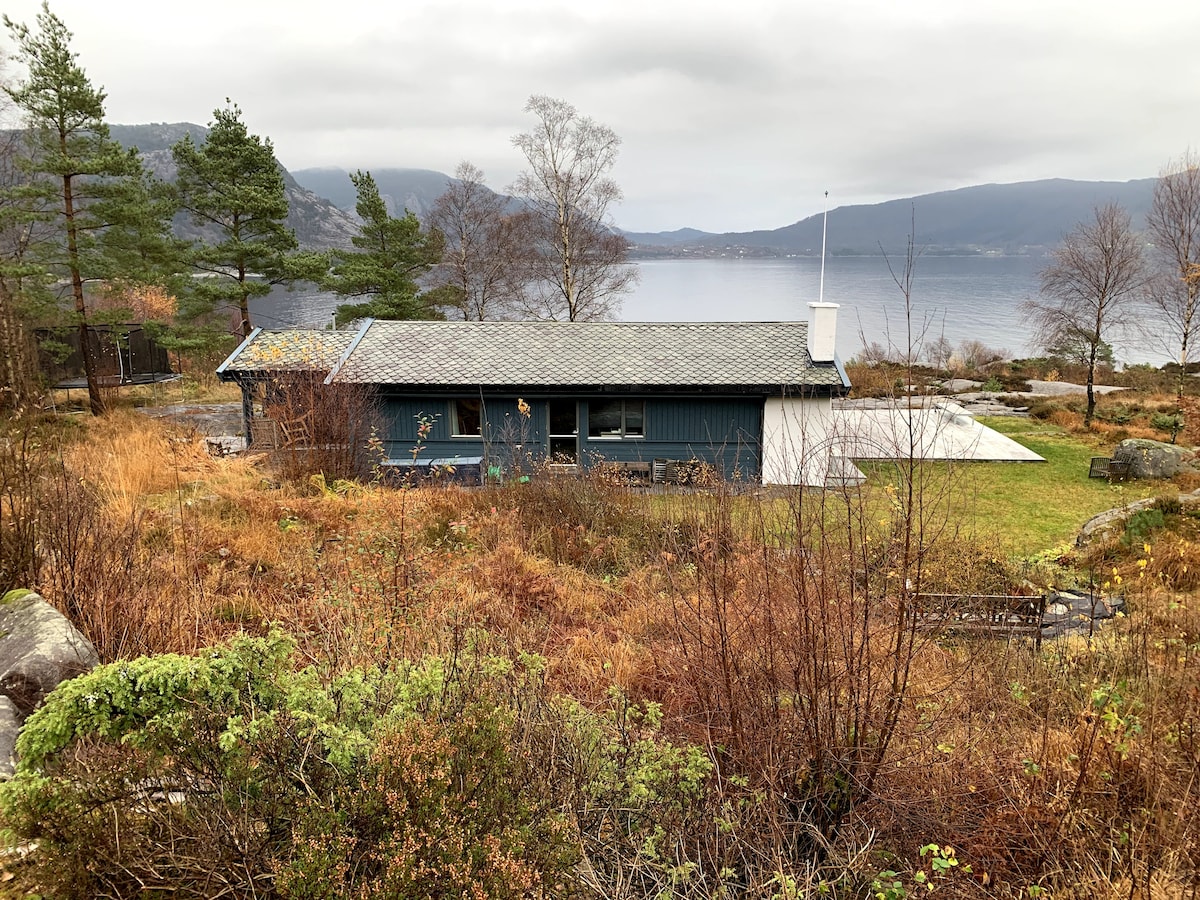 The cabin is nestled within a natural landscape, featuring greenery and rocky terrain. A serene fjord view is visible in the background. The structure is designed with a sloped roof and large windows, allowing for natural light and scenic views of the surroundings.