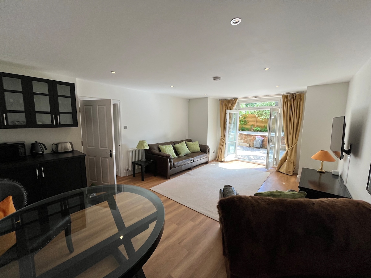 A bright living area features two comfortable sofas, accentuated by green cushions. Natural light floods the space through large glass doors, leading to a serene patio garden. A dining table and black cabinetry are included, creating a harmonious gathering zone.