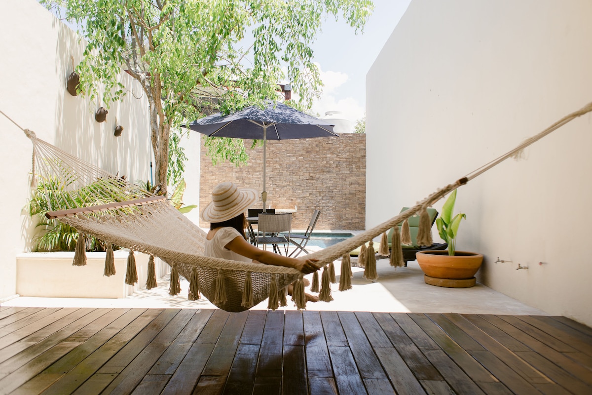 A hammock suspended between two points invites relaxation, with a shaded outdoor dining area in the background. A pool is visible further back, surrounded by a smooth stone wall and leafy plants, contributing to a serene outdoor setting.