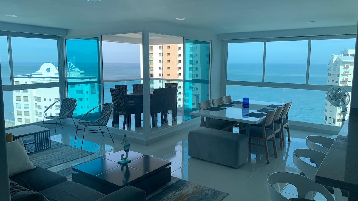 A spacious living area features large windows offering an unobstructed view of the ocean. Comfortable seating is arranged around a coffee table, and a dining table set for several is nearby. Natural light fills the space, complemented by a modern aesthetic.