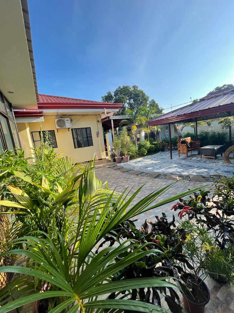 A spacious outdoor area is surrounded by various plants, offering a serene environment. The space features a paved floor and includes seating arrangements, creating an inviting spot for relaxation. A yellow building and shaded structure are visible in the background beneath a clear blue sky.