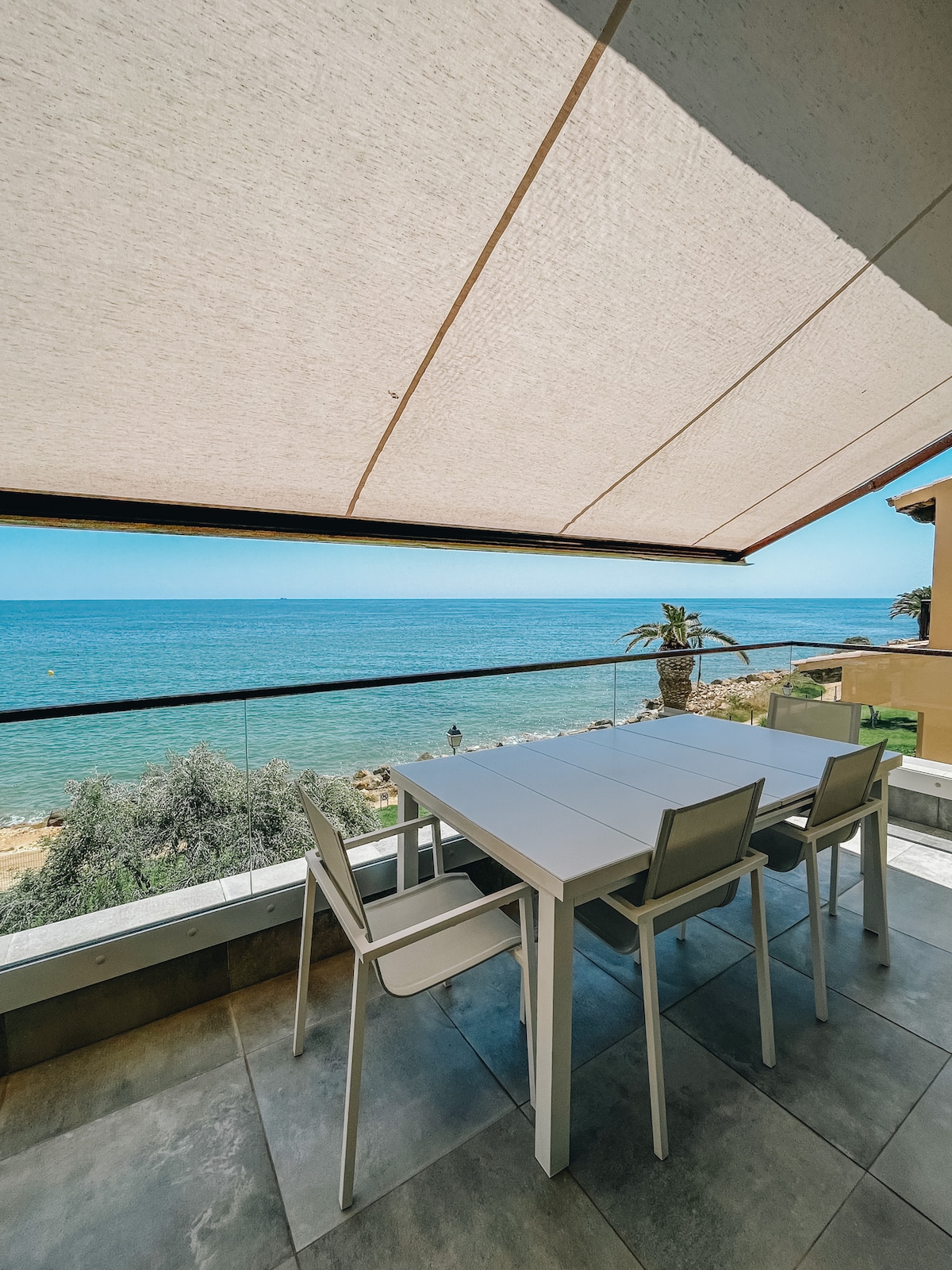 An outdoor dining area is presented with a sleek table and four chairs. A large awning provides shade while offering views of the clear blue sea and sky. The surroundings feature lush greenery and a pathway that leads towards the water.