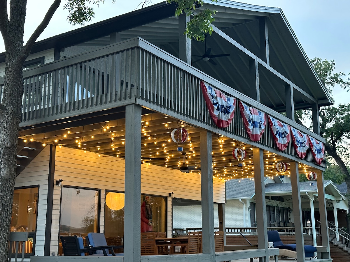 A spacious two-story deck features soft lighting and decorative bunting along the railing. Outdoor seating is arranged for relaxation and dining, with a view of the home's large glass windows that provide a glimpse of the interior.