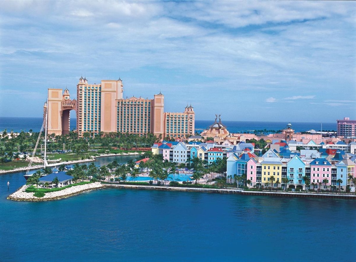 A vibrant view of the Harborside Resort at Atlantis is captured, showcasing the colorful villas along the waterfront. The tall resort towers rise in the background against a cloudy sky, while the serene blue water reflects the surrounding architecture and lush landscapes.