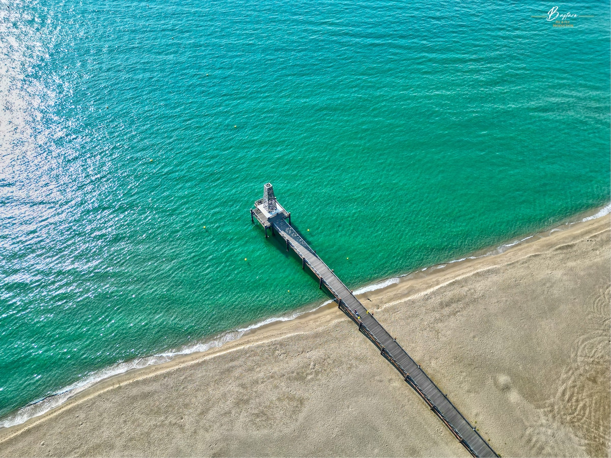 A wooden pier extends into the clear turquoise waters, framed by a sandy beach. The calm sea reflects sunlight, creating a serene environment. The shoreline is gentle, with minimal vegetation visible, promoting a tranquil coastal scene.