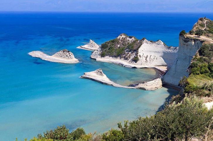 A coastal landscape displays clear turquoise waters against white rocky formations. Two small islets rise from the sea, while a cliff covered in greenery borders the shoreline. The scene offers a serene view of the horizon, with distant mountains visible across the water.