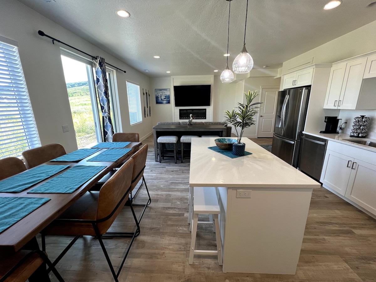 A light-filled kitchen and dining area features a modern dining table set for six and a spacious kitchen island with bar seating. Large glass doors open to a scenic view, and pendant lights hang above the island, enhancing the neutral color palette throughout the space.