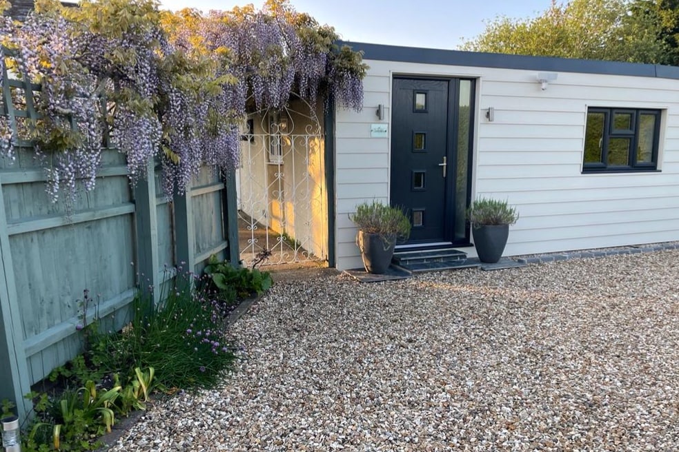 The entrance to the studio apartment is framed by vibrant purple wisteria cascading over a decorative gate. The exterior is clad in light-colored paneling, complemented by potted plants placed symmetrically at the entrance. Gravel pathways provide access to the front door.