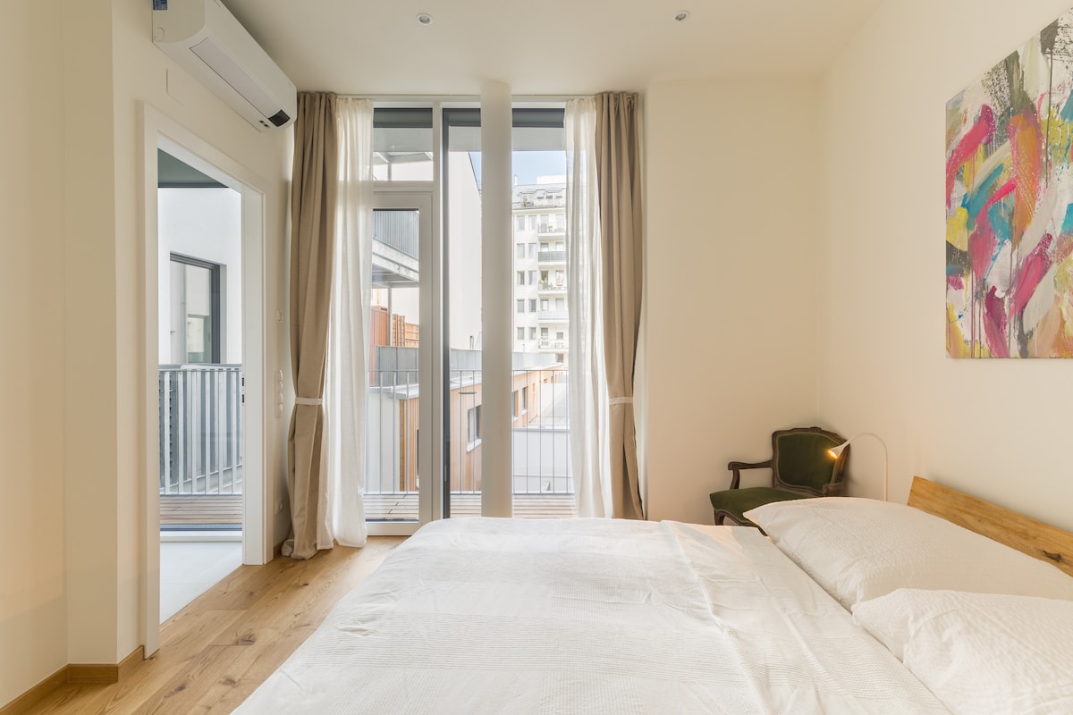 A bright bedroom features a spacious bed covered with crisp white linens. Large windows allow natural light to fill the room, with sheer curtains draping softly. A small green armchair is positioned nearby, and a colorful piece of artwork adorns the wall.