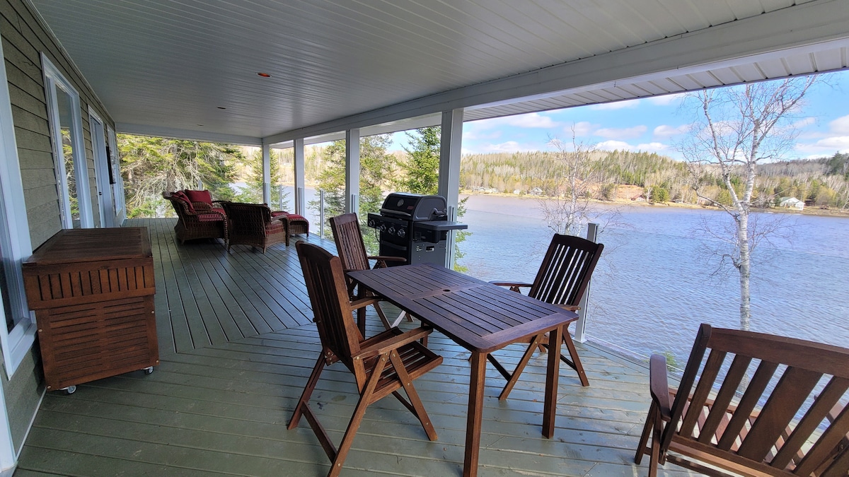 A covered outdoor deck features a wooden dining table surrounded by four chairs and a barbecue grill. Two lounge chairs are positioned nearby, offering a scenic view of the Tobique River and surrounding trees under a clear blue sky.