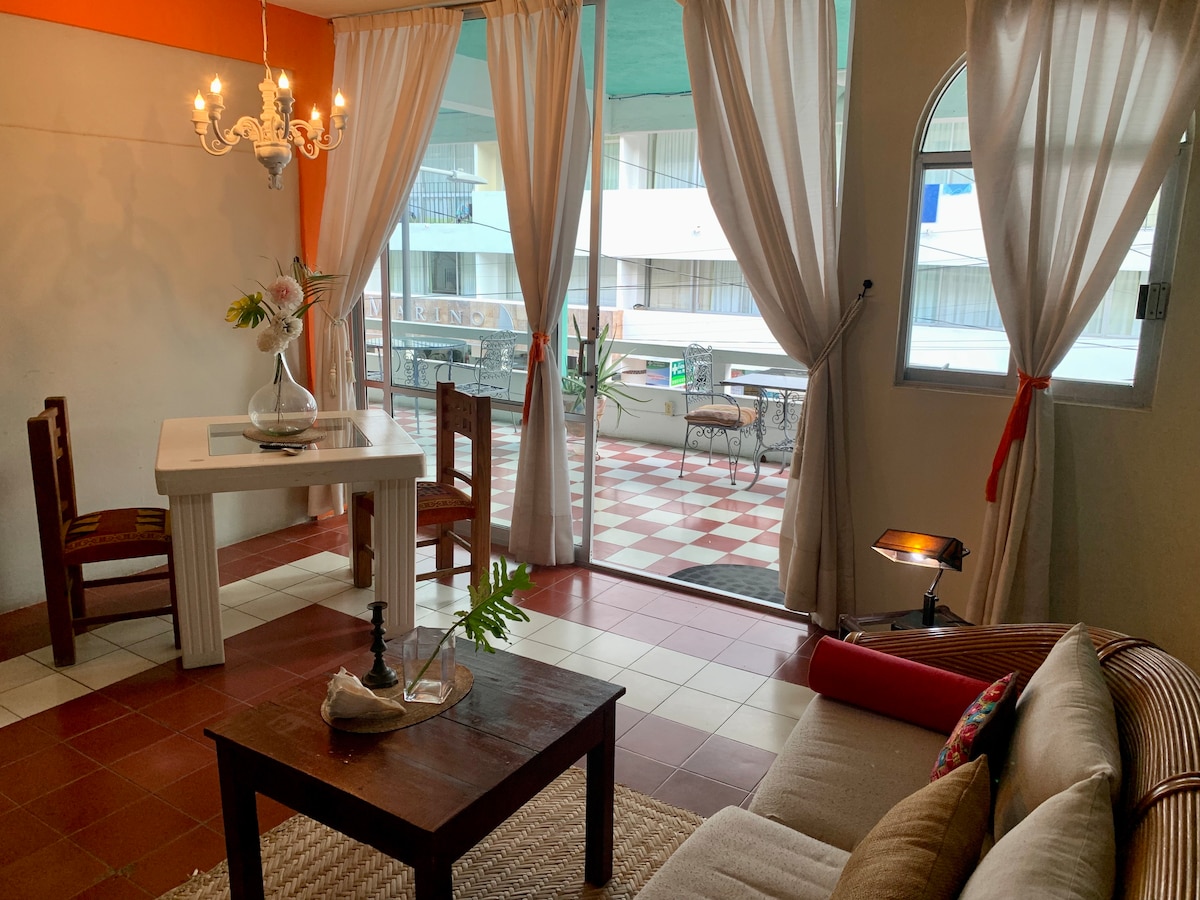 An inviting living space displays a cozy sofa and a rustic wooden coffee table at its center. A dining table is positioned nearby, surrounded by wooden chairs. Sheer curtains frame the windows, allowing natural light to fill the room, leading to a balcony with a checkerboard floor visible beyond.