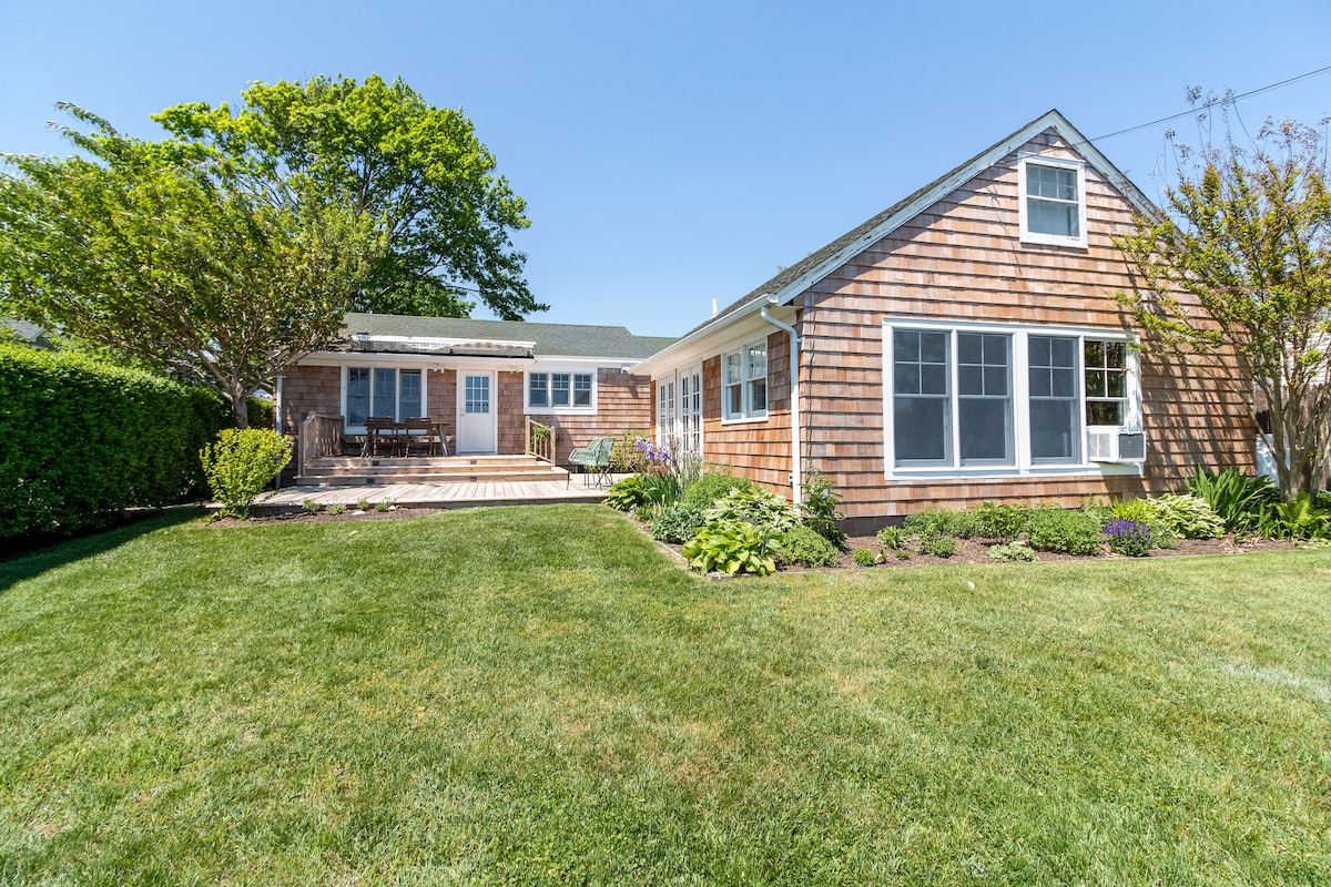 The exterior of the cottage is highlighted by a well-maintained yard featuring a lush green lawn. A patio area with seating is visible, surrounded by flowering shrubs and trees. Multiple large windows offer a view into the inviting living spaces within.