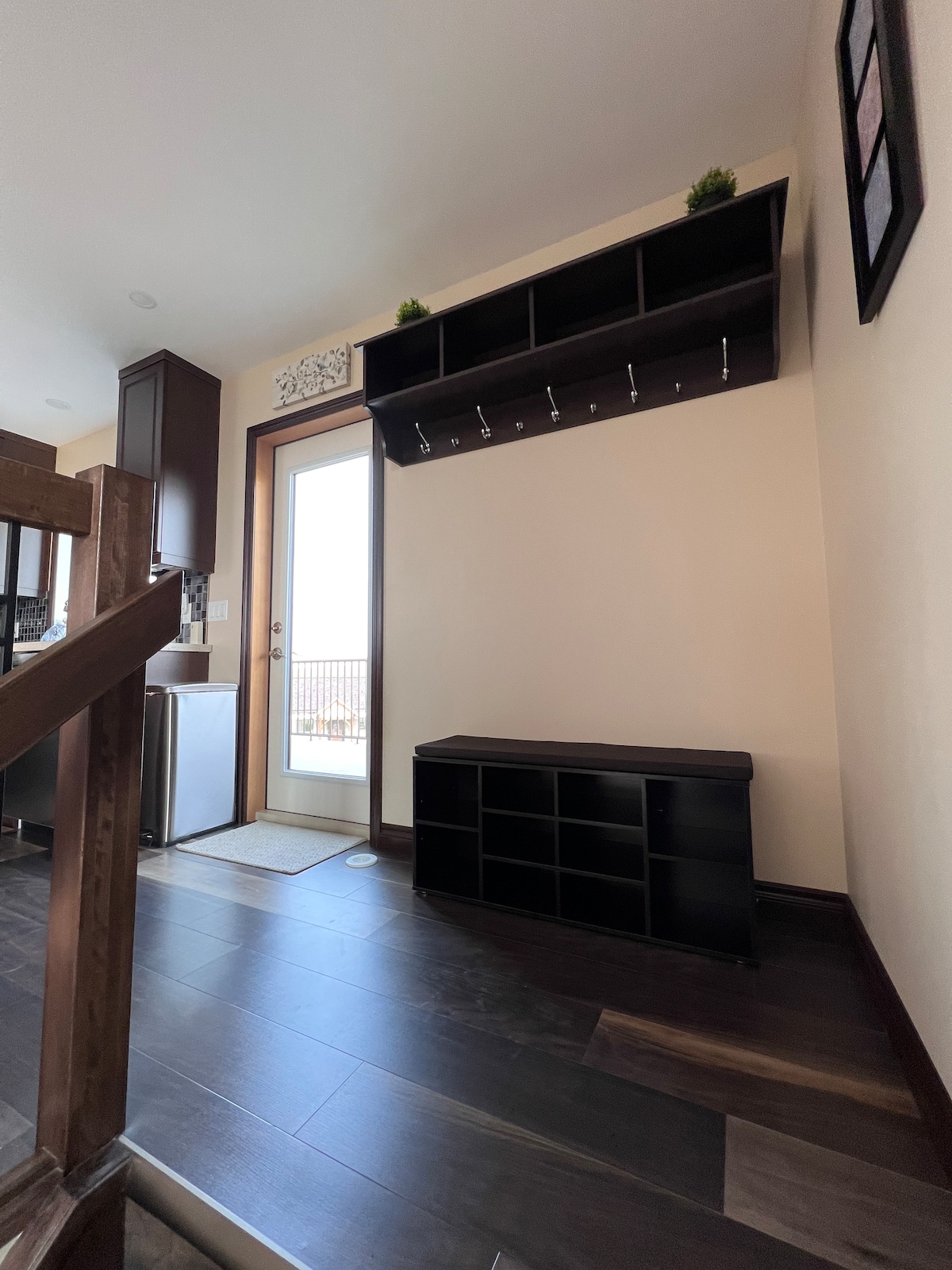 A well-organized entryway features a storage bench with cubbies and wall-mounted hooks above. Natural light is welcomed through the door leading to an outdoor space. Dark wood flooring adds warmth to the area, creating a functional and inviting entrance.