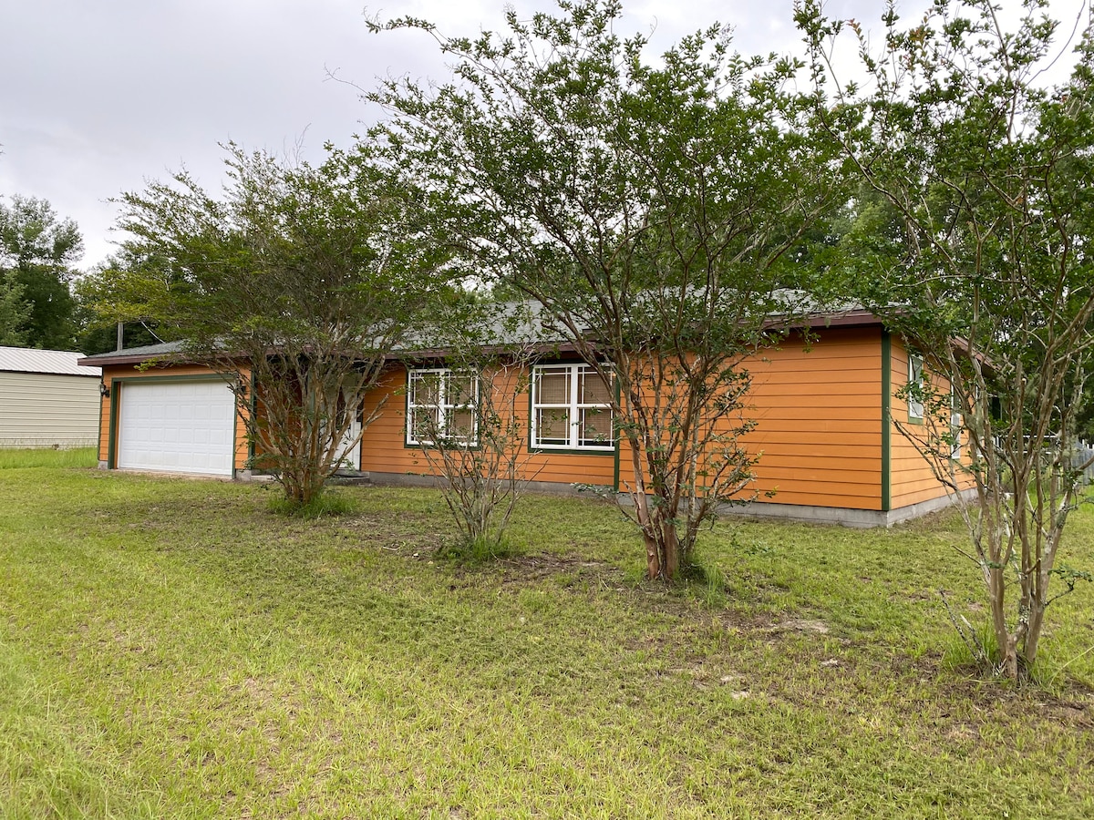 A single-story home features an inviting orange exterior contrasted by white trim. A garage door is visible, alongside multiple windows framed by greenery. A well-maintained lawn provides ample space, with patches of grass surrounding the home and small shrubs scattered throughout.