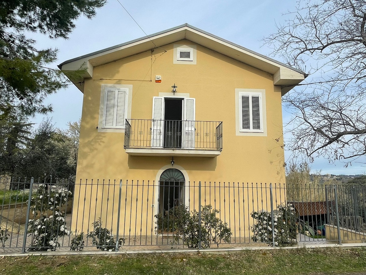 A two-story yellow house is presented, featuring white shutters and a front balcony. A decorative iron fence surrounds the property, with green shrubs visible along the base. The entrance door is centrally located, adding charm to the inviting façade.