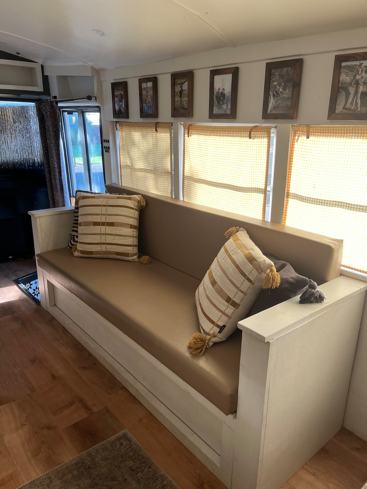 A cozy seating area features a long couch adorned with decorative pillows, enhancing comfort. Natural light filters through woven shades, illuminating the space. Walls are adorned with framed photographs, adding a personal touch to the interior design.