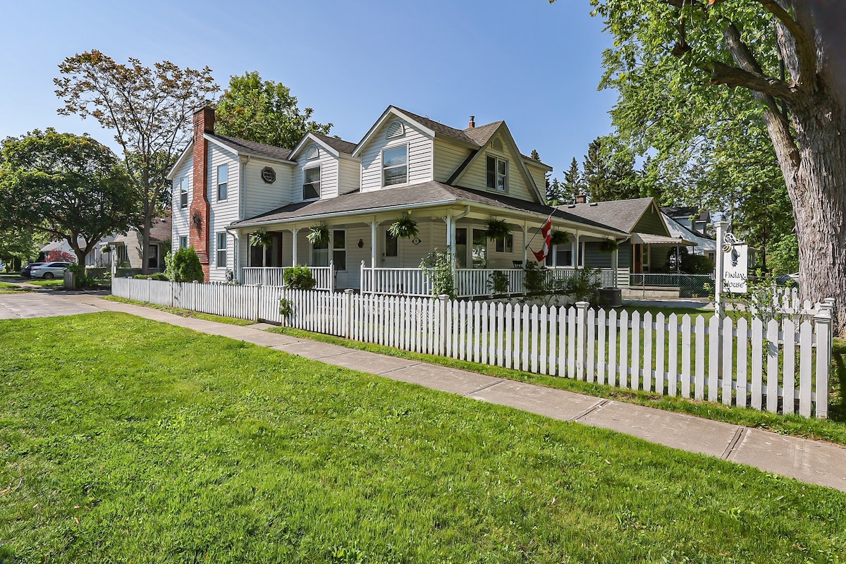 A charming house is set back from the street, featuring a welcoming front porch with white railings and hanging plants. The property is surrounded by a well-maintained green lawn and a white picket fence, offering a delightful view of the neighborhood.