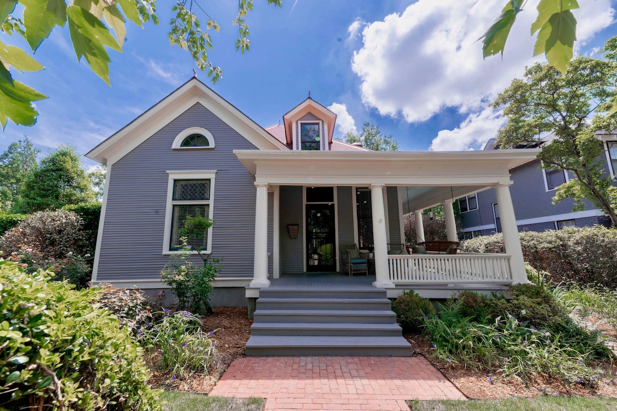 The charming exterior of a historical home is captured, showcasing a large front porch with steps leading to the entrance. Lush greenery and colorful foliage frame the house, which features distinctive architectural details and a unique roofline against a backdrop of bright blue skies.