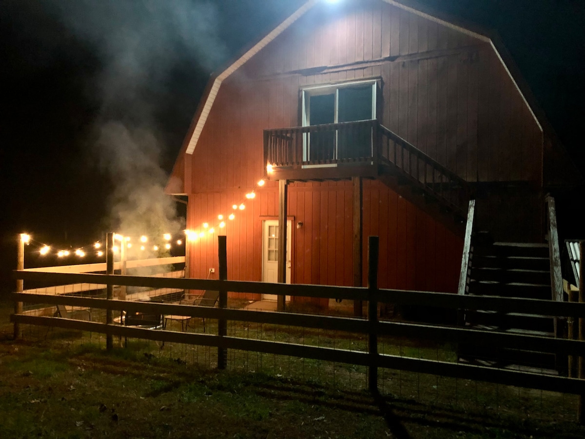 A warmly lit barn exterior is visible at night, featuring a second-floor balcony and string lights that create a welcoming ambiance. A wooden fence surrounds the property, and smoke can be seen rising from a fire pit in front, enhancing the serene environment.