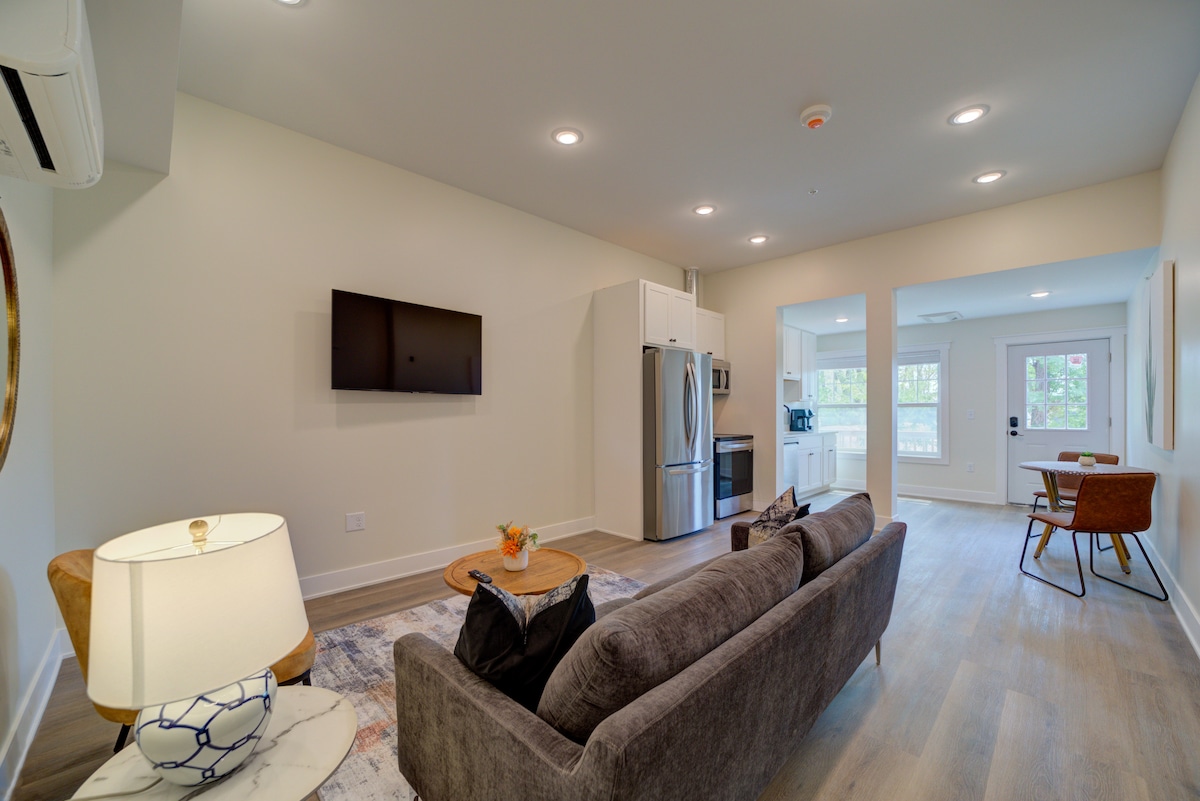 A cozy living area is highlighted by a comfortable sofa and a round coffee table. A mounted flat-screen television is positioned on a light wall, while natural light flows in from the entryway. A dining table is set for two against the far wall.