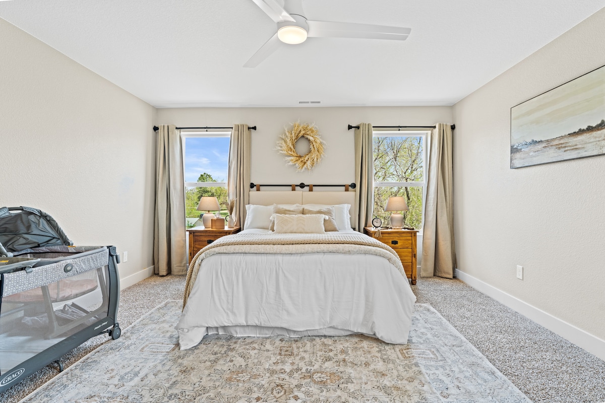 The master bedroom offers a spacious layout featuring a comfortable bed with neutral bedding. Two bedside tables with lamps are placed beside the bed, while large windows allow natural light to brighten the room. A Pack ‘N Play is positioned in the corner.