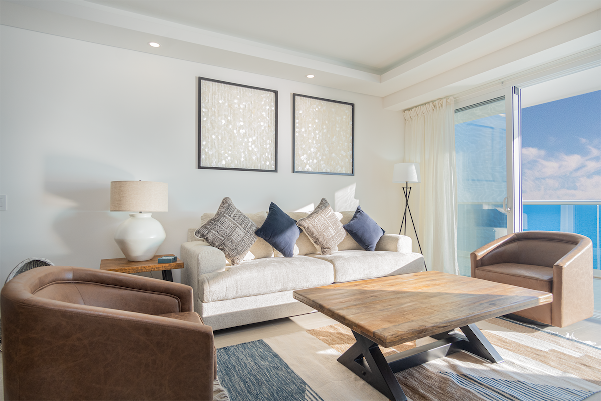 A bright living area features a spacious cream sofa adorned with decorative pillows, complemented by two brown armchairs. A wooden coffee table is centered on a stylish area rug. Abstract artwork hangs on the wall, and a sliding glass door provides scenic ocean views.