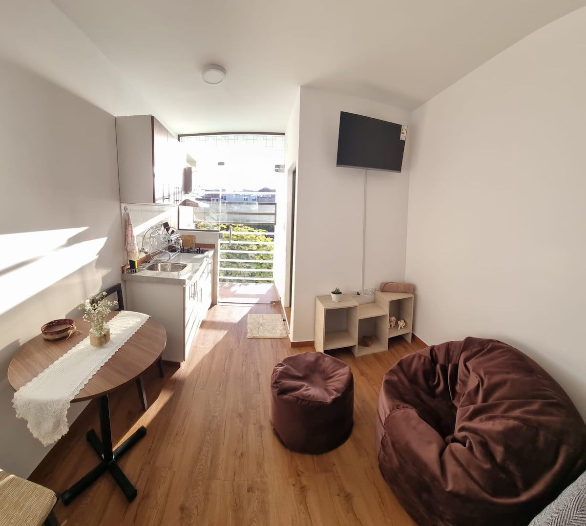 A comfortable living area features a small round table next to a large bean bag, with a kitchenette visible in the background. Natural light streams through the sliding door, highlighting the modern design. A television is mounted on the wall opposite the seating area.