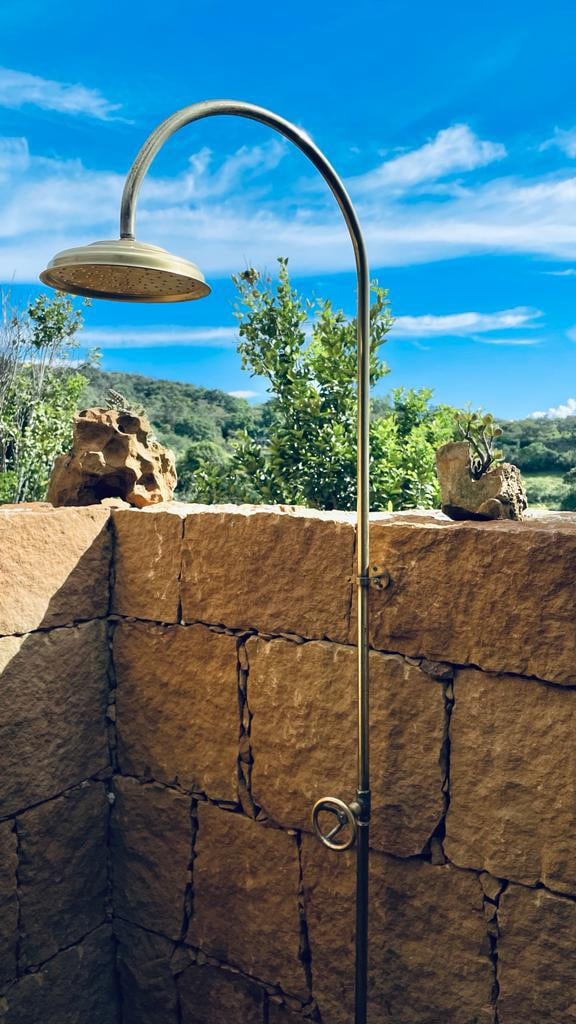 An outdoor shower is positioned against a rustic stone wall, featuring a curved brass showerhead. Lush greenery is visible beyond the wall, with the sky showcasing a bright blue hue and scattered clouds.