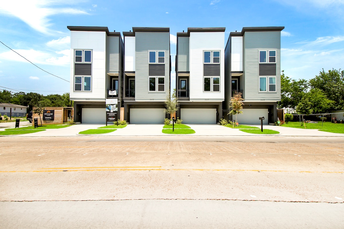 A modern three-story building features a sleek design with multiple large windows and a ground-level parking area. The front yard is neatly landscaped, showcasing green grass and small trees. The facade has contrasting colors, giving it a contemporary appearance.