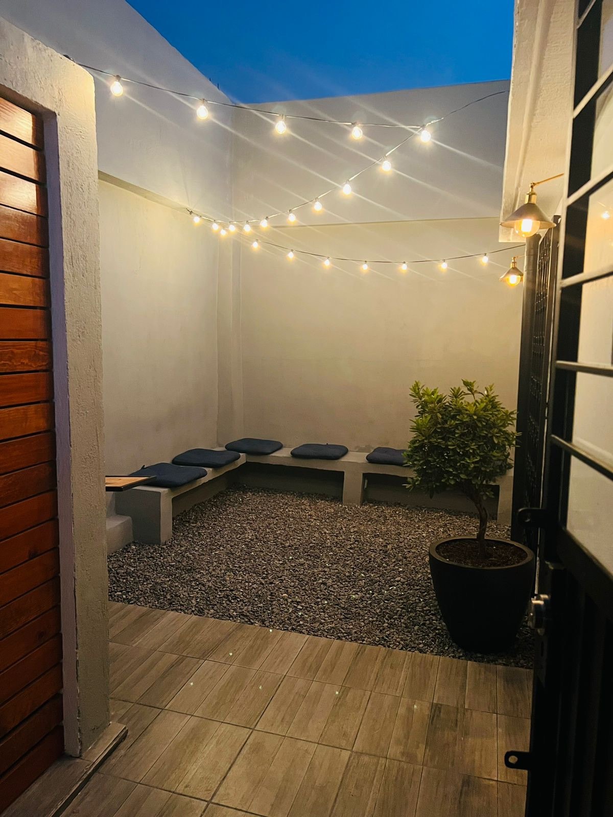 A tranquil patio area features comfortable stone seating covered with blue cushions, surrounded by gravel. Soft lighting is provided by string lights overhead, creating a serene environment. A small potted plant adds a touch of greenery, enhancing the inviting atmosphere of the space.